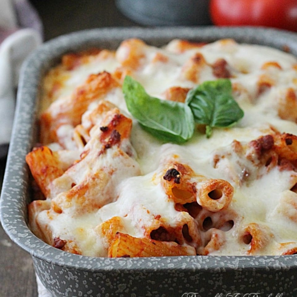 Baked pasta topped with melted mozzarella, browned cheese, and fresh basil in a speckled ceramic dish.