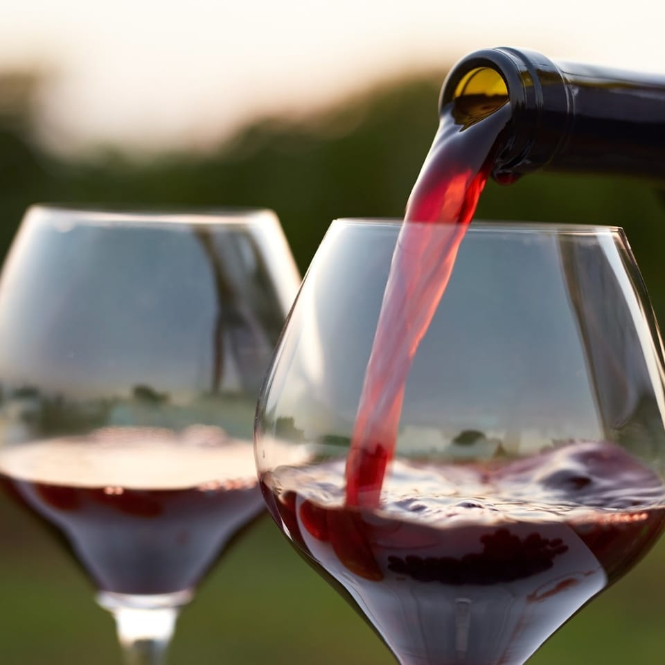 Red wine pours from a bottle into a glass, with a second filled wine glass against a softly blurred outdoor backdrop.