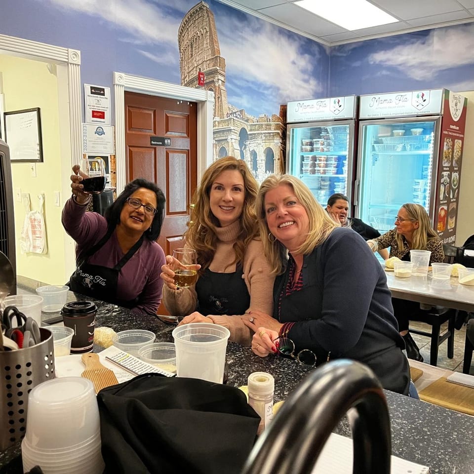 Three women smile and raise wine glasses inside Mama Mia Italian restaurant, surrounded by food containers and Roman-themed decor.