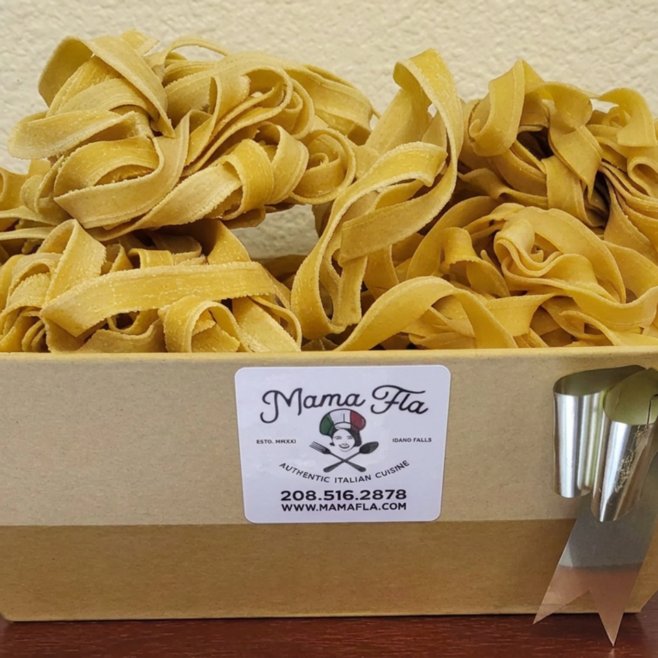 Fresh tagliatelle pasta fills a cardboard gift box labeled Mama Fla Authentic Italian Cuisine, accented with a silver ribbon.