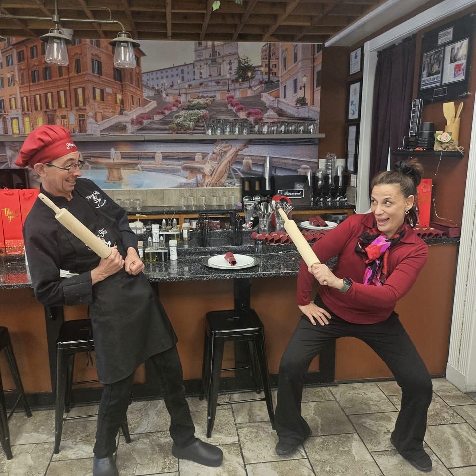 Two restaurant staff playfully pose with rolling pins at a bar beneath a mural of Rome’s Spanish Steps.