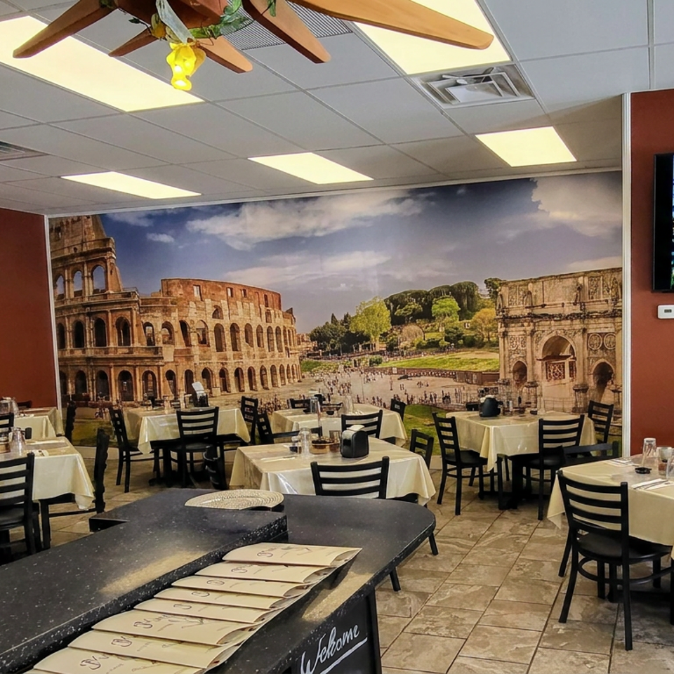 Italian restaurant dining room with white tablecloths, black chairs, Colosseum mural, and mountain landscape displayed on a television.