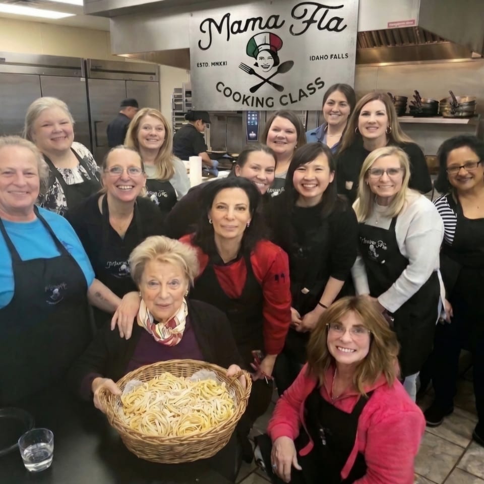 Group of cooking class participants posing in a commercial kitchen beneath a Mama Fla Cooking Class sign, holding a basket of fresh pasta.