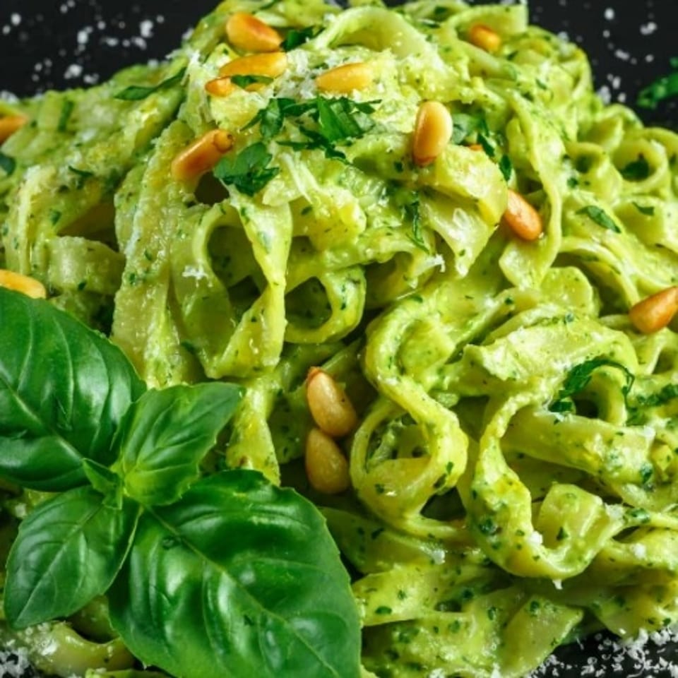 Creamy green pesto pasta topped with pine nuts and fresh basil leaves on a black plate.