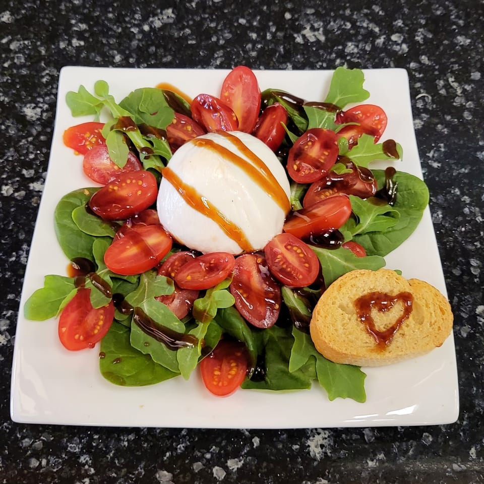 Burrata salad with mixed greens, halved tomatoes, balsamic glaze, and toasted bread on a white plate.