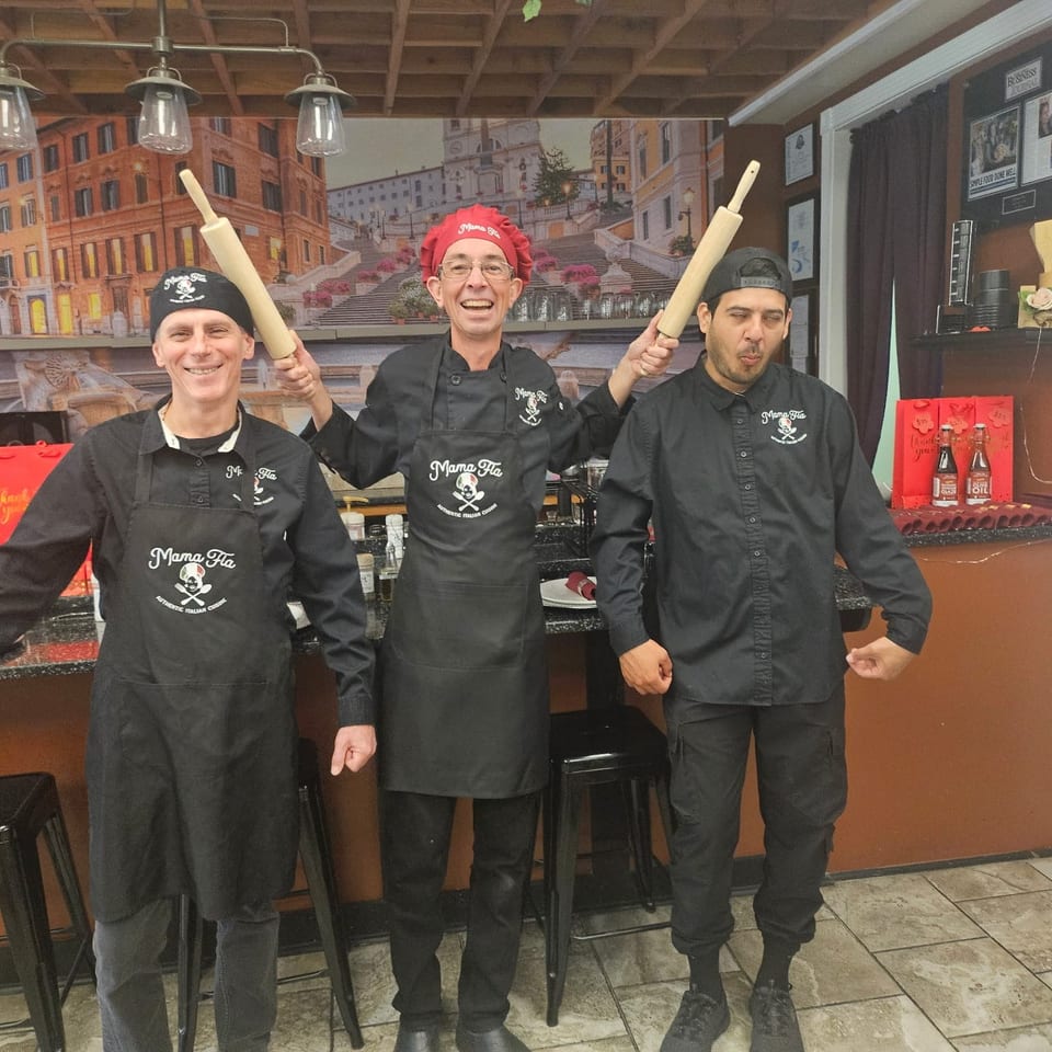 Three smiling Mama Mia restaurant staff members pose in black uniforms, with the chef holding two rolling pins in an Italian-themed dining area.